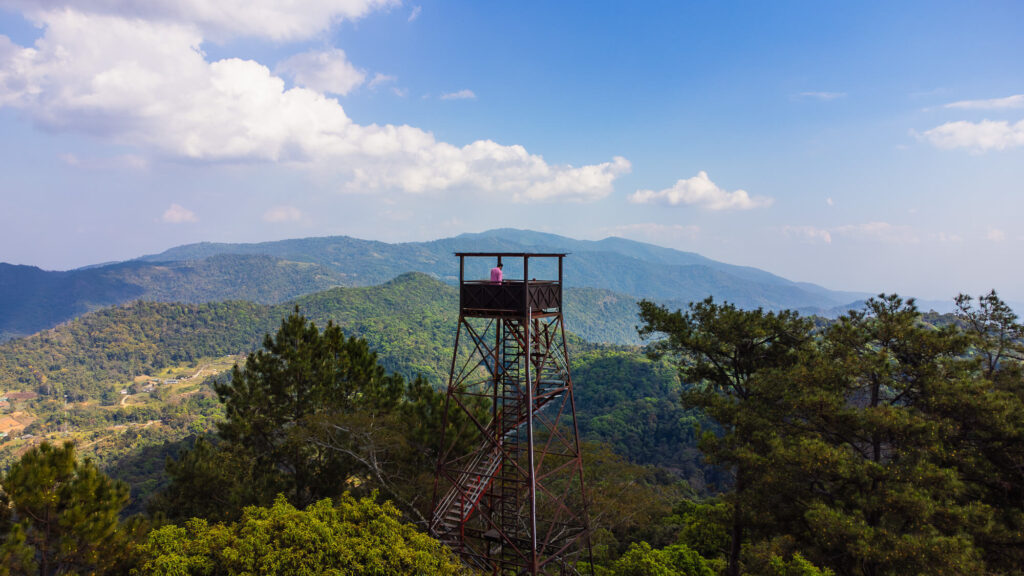 A drone shot of a traveler standing on the Buak Chan Fire Observation Tower, the best hidden Samoeng Loop stop near Chiang Mai.