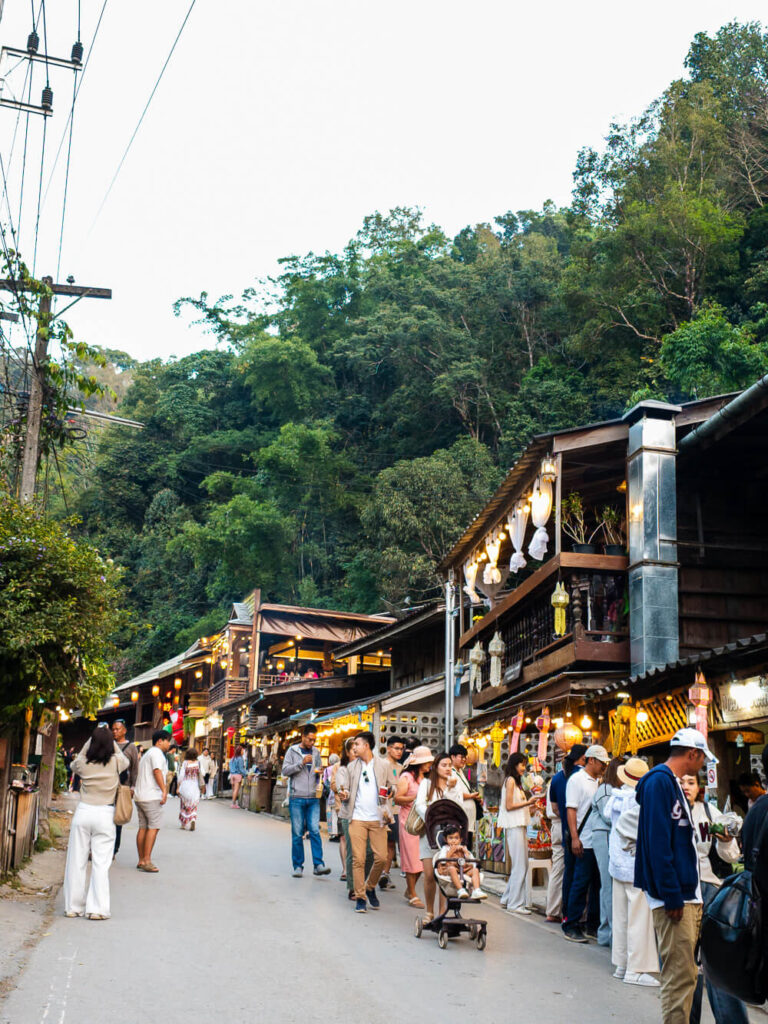 A narrow road in Mae Kampong Village packed with tourists and parked scooters during a busy Saturday.