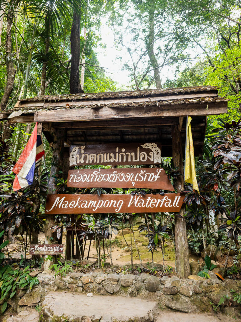 A wooden sign marking the entrance to the Mae Kampong Waterfall trail in the forest.