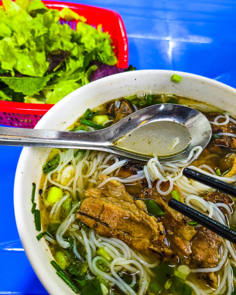 A bowl of authentic bún chả with grilled pork, noodles, and fresh herbs served on a plastic table in Hanoi.