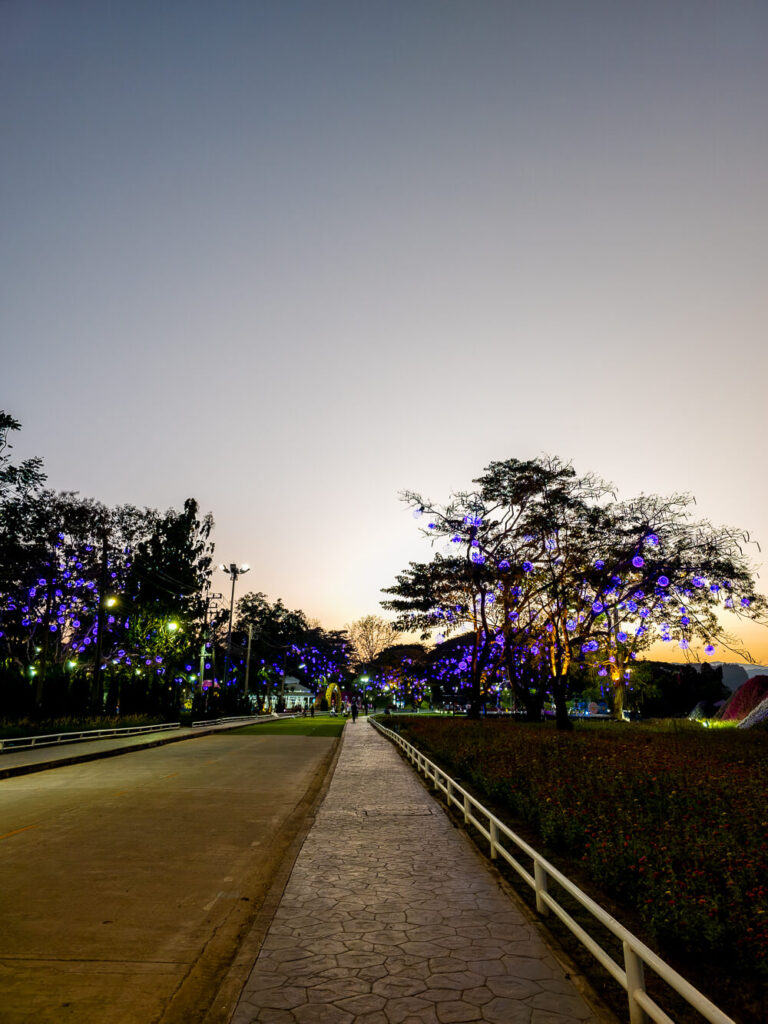 A wide shot of the Kok River in Chiang Rai at sunset with warm lights glowing from riverside stalls and people relaxing on the sand.