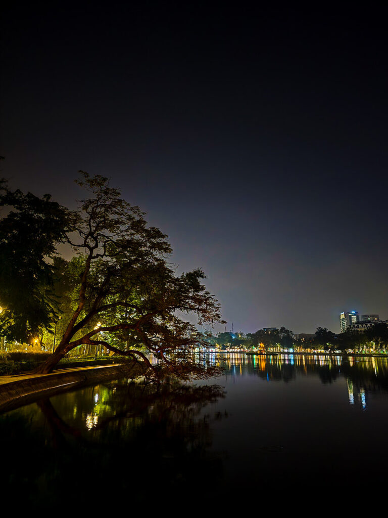 A large illuminated heritage tree leaning over the dark water of Hoàn Kiếm Lake in Hanoi at night
