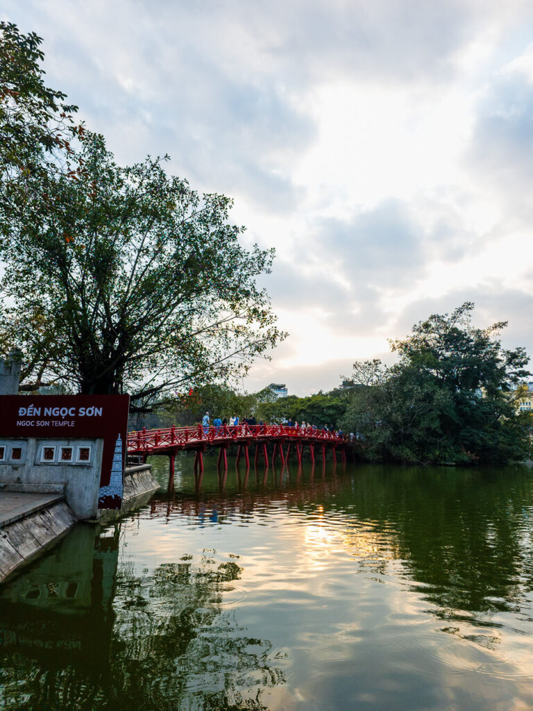 Daylight view of Ngoc Son Temple sitting on a small island in Hoan Kiem Lake surrounded by lush green trees.