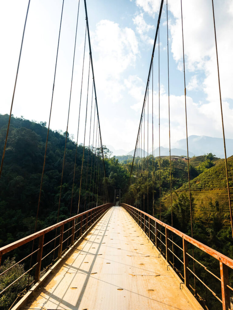 A wooden and steel suspension bridge crossing a river in the foggy, terraced mountains near Sapa, Northern Vietnam.