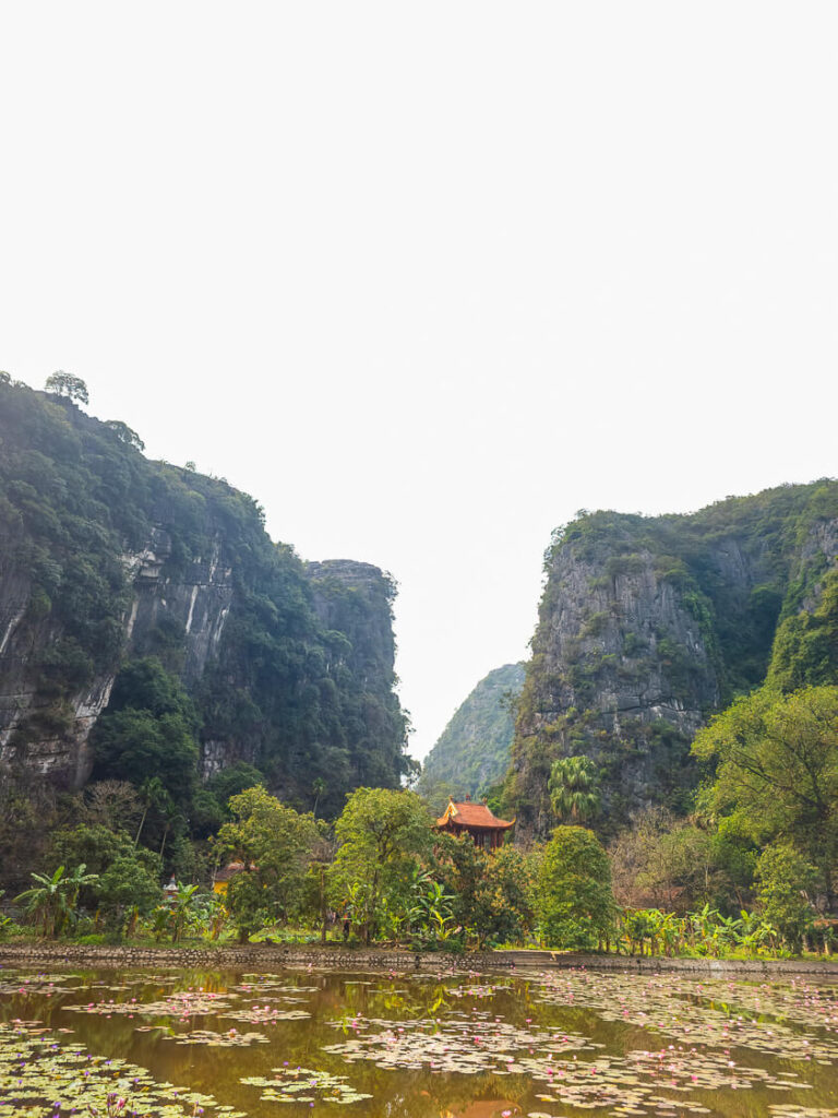 The historic stone bridge and arched entrance gate of the ancient Bich Dong Pagoda surrounded by a green lotus pond and limestone mountains during the day.