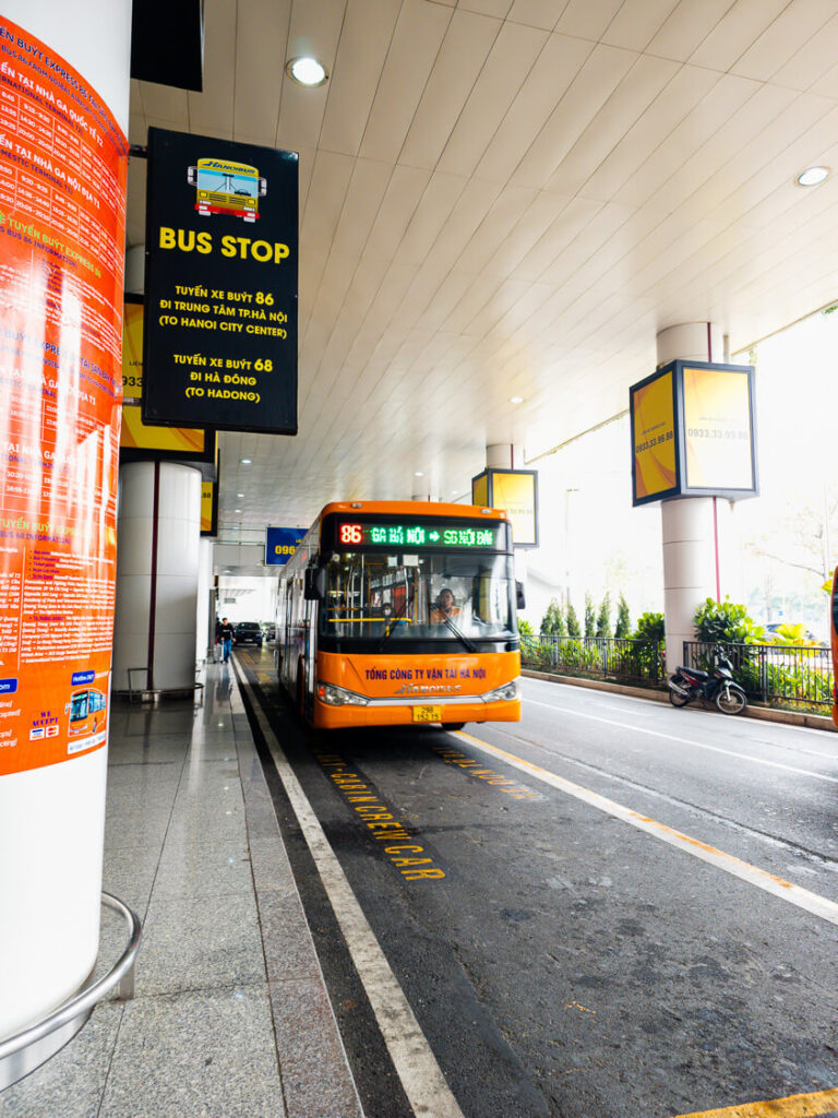 The orange and white Hanoi Airport Bus 86 pulling up to the curb at Noi Bai International Airport to pick up passengers.