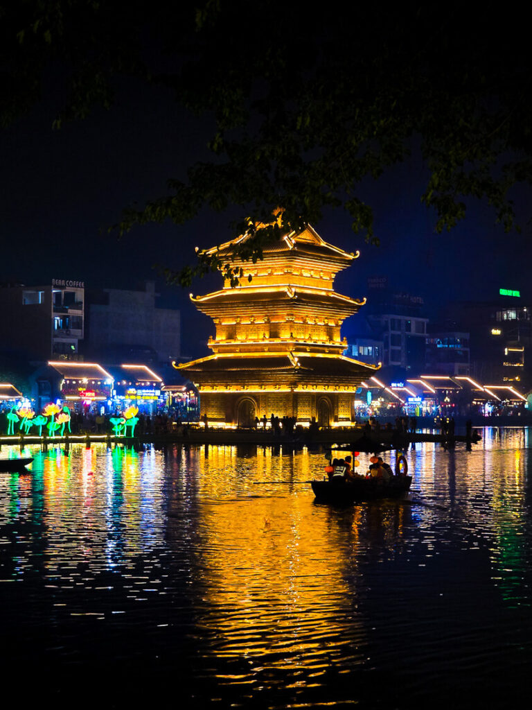 A traditional wooden boat docked in front of the illuminated Thap Tu An temple reflecting on the water at night.