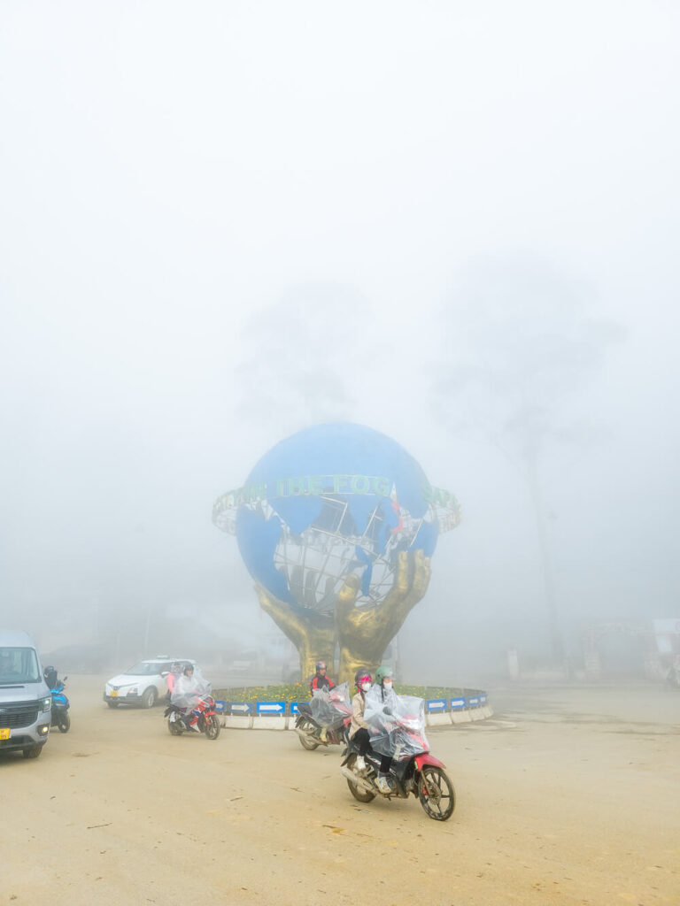 The stone globe held by hands statue near the sapa church covered in heavy fog with scooters parked in the front