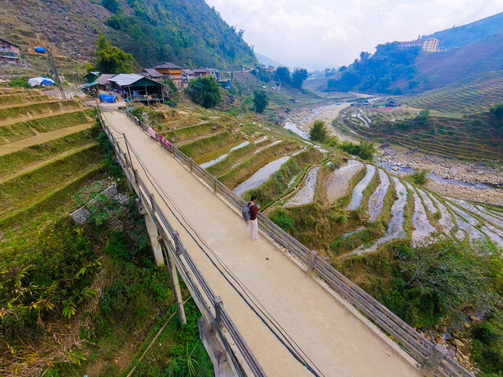 High-angle drone shot of a traveler standing on a concrete bridge near a mountain village in Sapa, Vietnam.