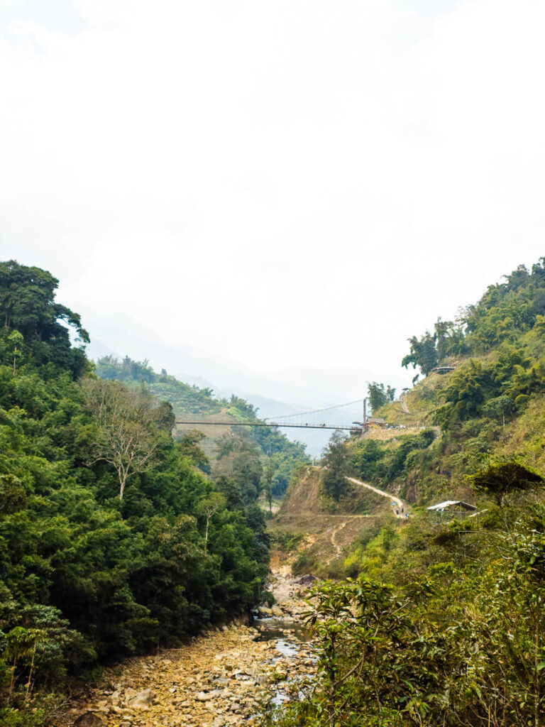 Wide panoramic view of a rustic suspension bridge spanning a river in the Muong Hoa Valley, Sapa, surrounded by mountains.