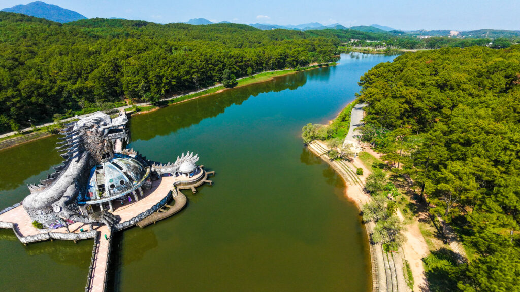 An aerial drone shot showing the massive stone dragon at Ho Thuy Tien water park sitting in a drained lagoon surrounded by pine forest.