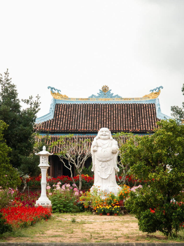 A large ornate statue of a laughing Buddha sitting peacefully outside a traditional Buddhist temple in Vietnam.