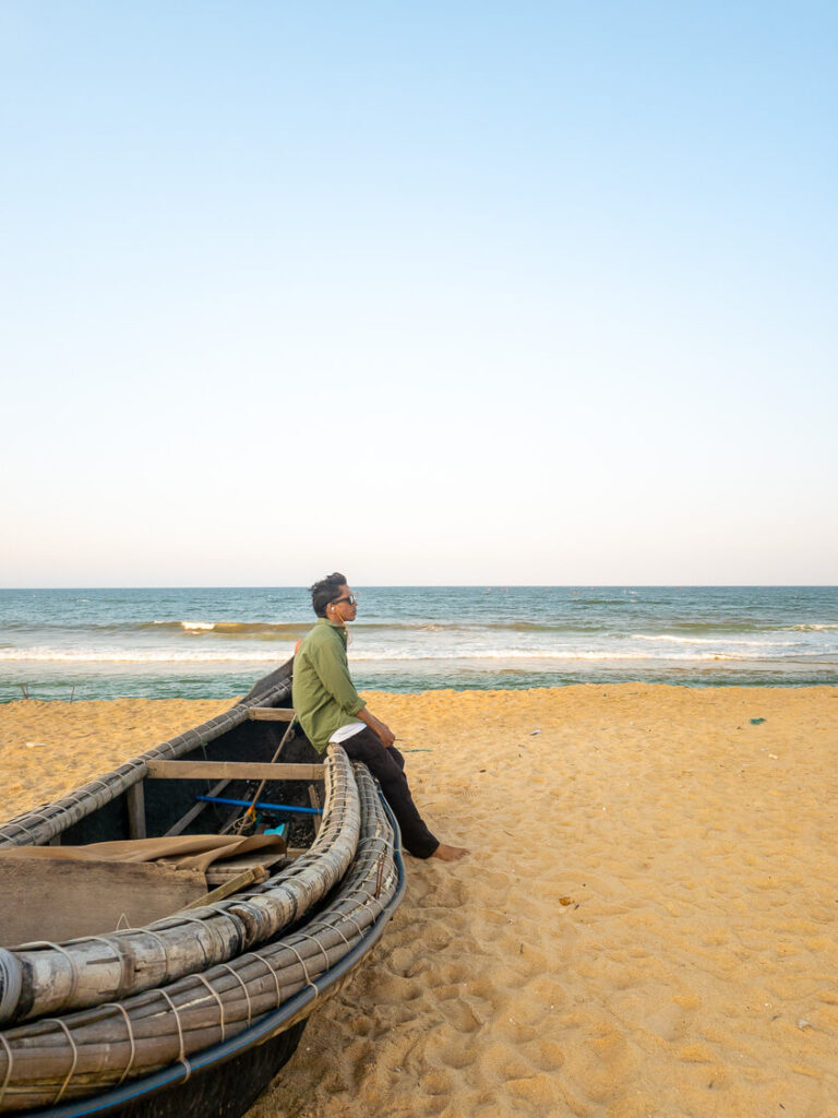A traveler sitting quietly on a traditional wooden fishing boat resting on the sand at Thuan An Beach, an offbeat coastal spot near Hue.