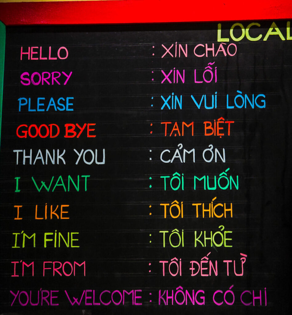 A wooden board displaying basic English to Vietnamese translations for common daily phrases like hello and how are you.