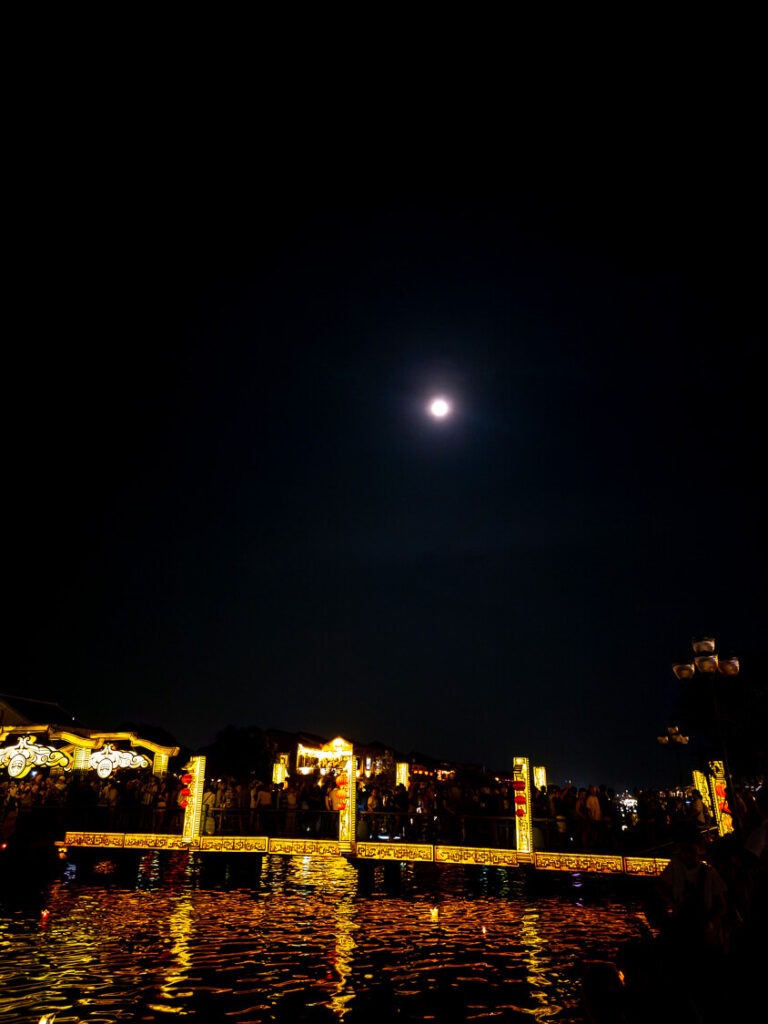 A night shot of the illuminated bridge connecting two islands near the famous boat lantern rides in Hoi An, glowing under a bright moon.