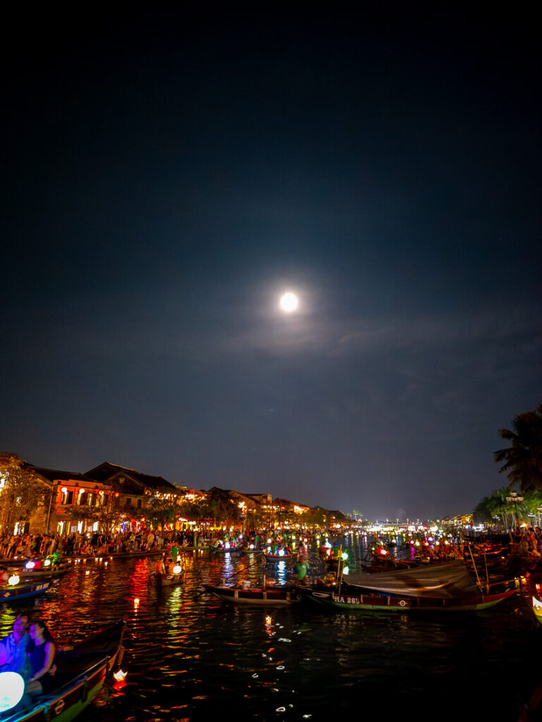 Traditional wooden boats illuminated by colorful paper lanterns floating down the Thu Bon River in Hoi An under a bright moon.