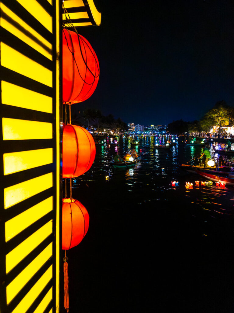 An elevated nighttime view from a decorated bridge looking down at colorful lantern-lit boats drifting along the Thu Bon River in Hoi An.