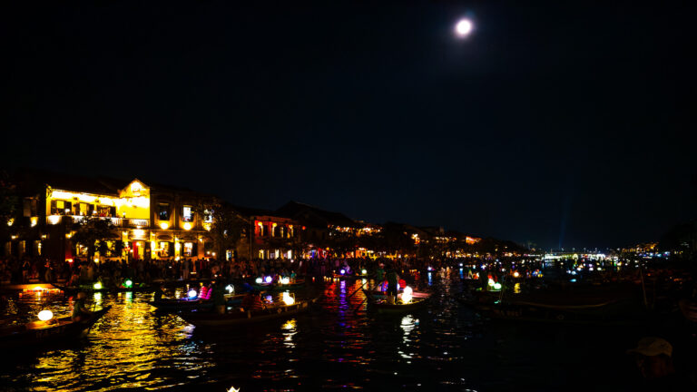 Traditional wooden boats floating on the Thu Bon River during a Hoi An river boat ride at night, illuminated by colorful lanterns and a glowing moon.