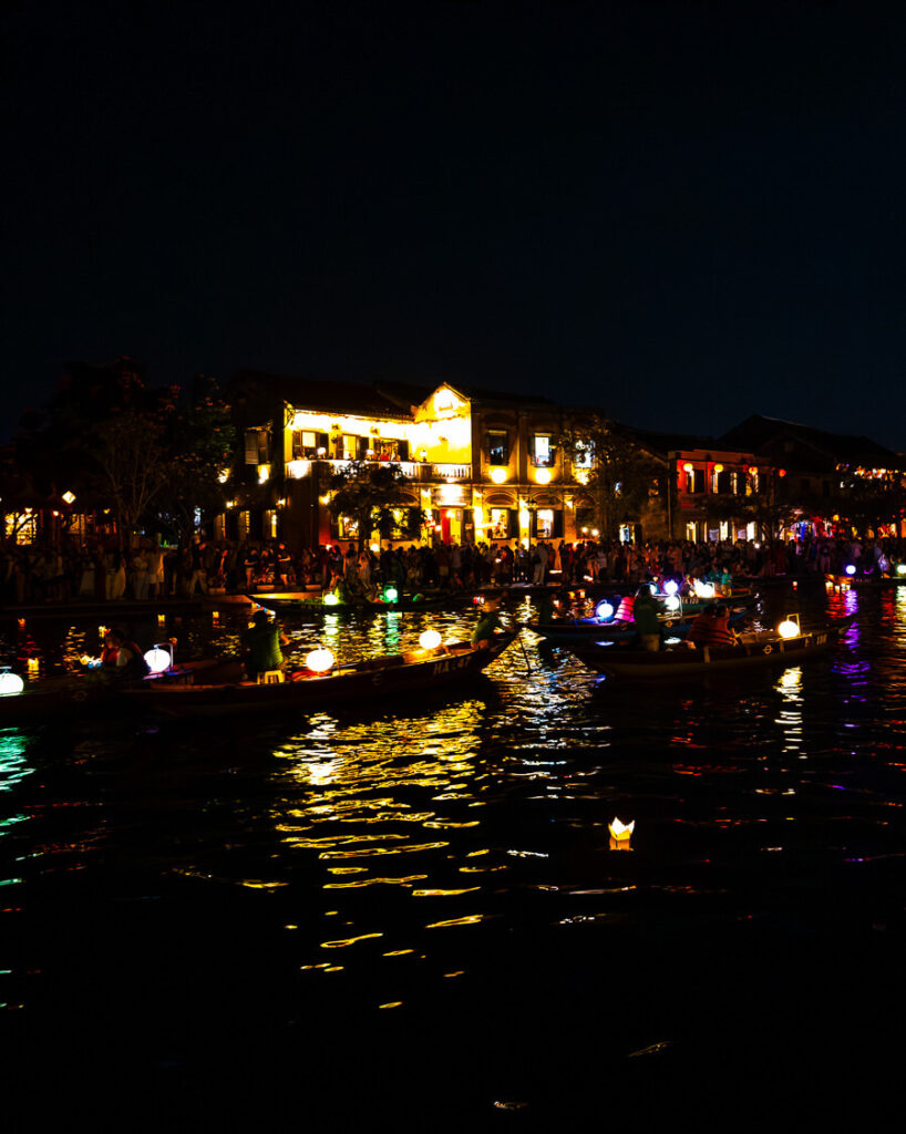 Several traditional wooden hand-rowed boats glowing with paper lanterns navigating the calm waters of the river at twilight.