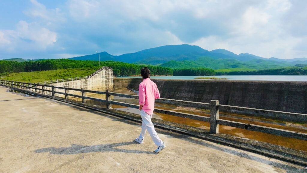 Vishal walking across a concrete bridge with the calm waters of Khe Tan Reservoir in the background.
