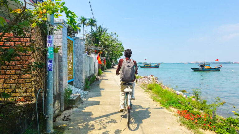 A traveler cycling along the waterfront on Cam Kim Island with traditional Vietnamese fishing boats docked in the background.