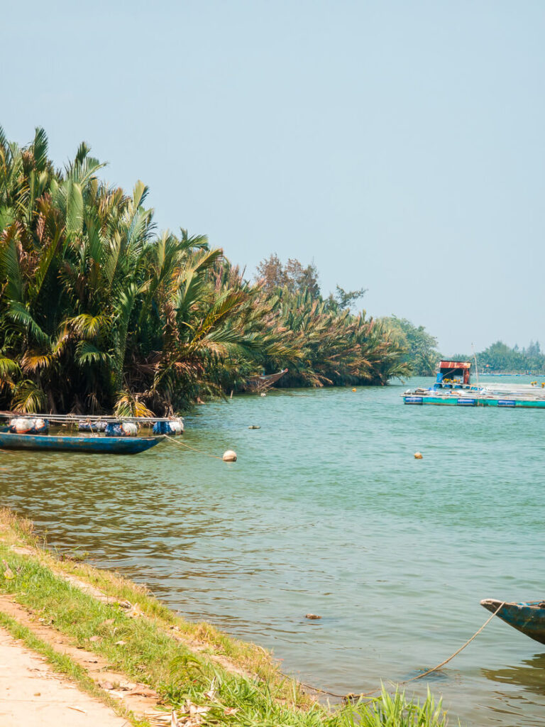 A view of a quiet riverbank lined with green trees and small fishing boats docked along the water.