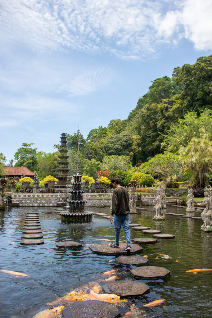 How to Plan a Trip to Bali 2026: The Ultimate Regional Travel Guide 7 A traveler walking across the traditional stone platforms in the water while surrounded by large golden koi fish at Tirta Gangga.